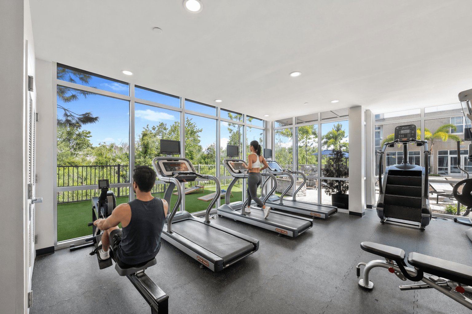A man is working out on a rowing machine in a spacious gym with a view of the outdoors.